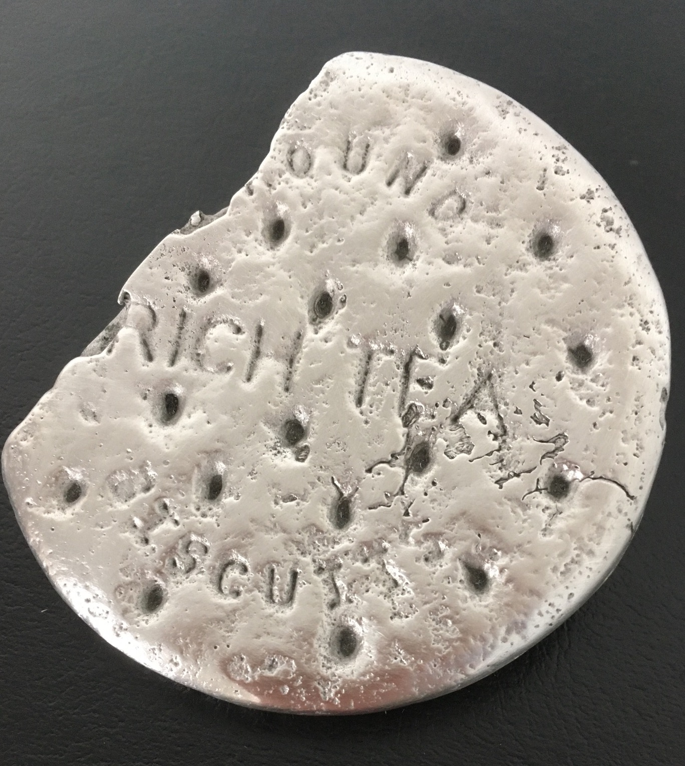 A silver-coloured metal sculpture of a broken round biscuit with the embossed text "OUND RICH TEA," displayed under a clear glass dome on a white base.