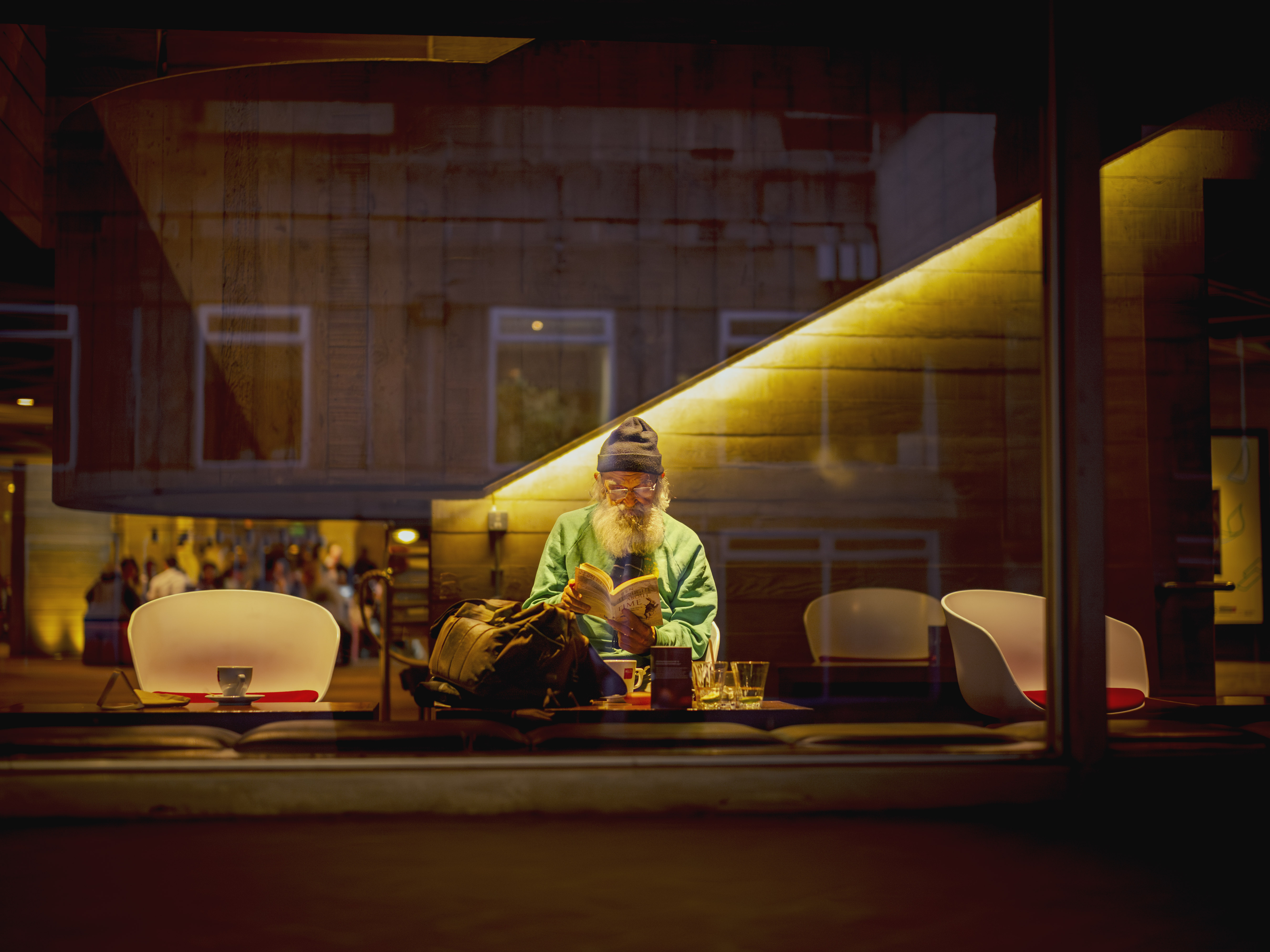 A photograph of an elderly man with a white beard reading a book at a theatre cafe table, with glasses and a backpack beside him, and blurred figures visible in the background.