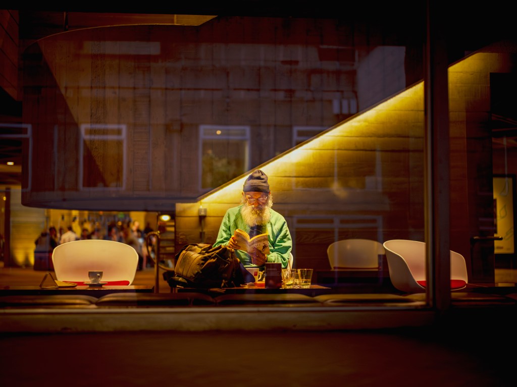 A photograph of an elderly man with a white beard reading a book at a theatre cafe table, with glasses and a backpack beside him, and blurred figures visible in the background.