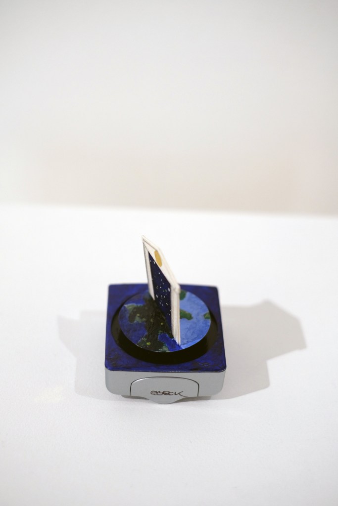 A miniature art installation which rotates on the face of a small battery-powered clock. A paper crafted classical facade depicts blue sky and clouds, set against a pink background, turns to reveal the night time view on the reverse side.