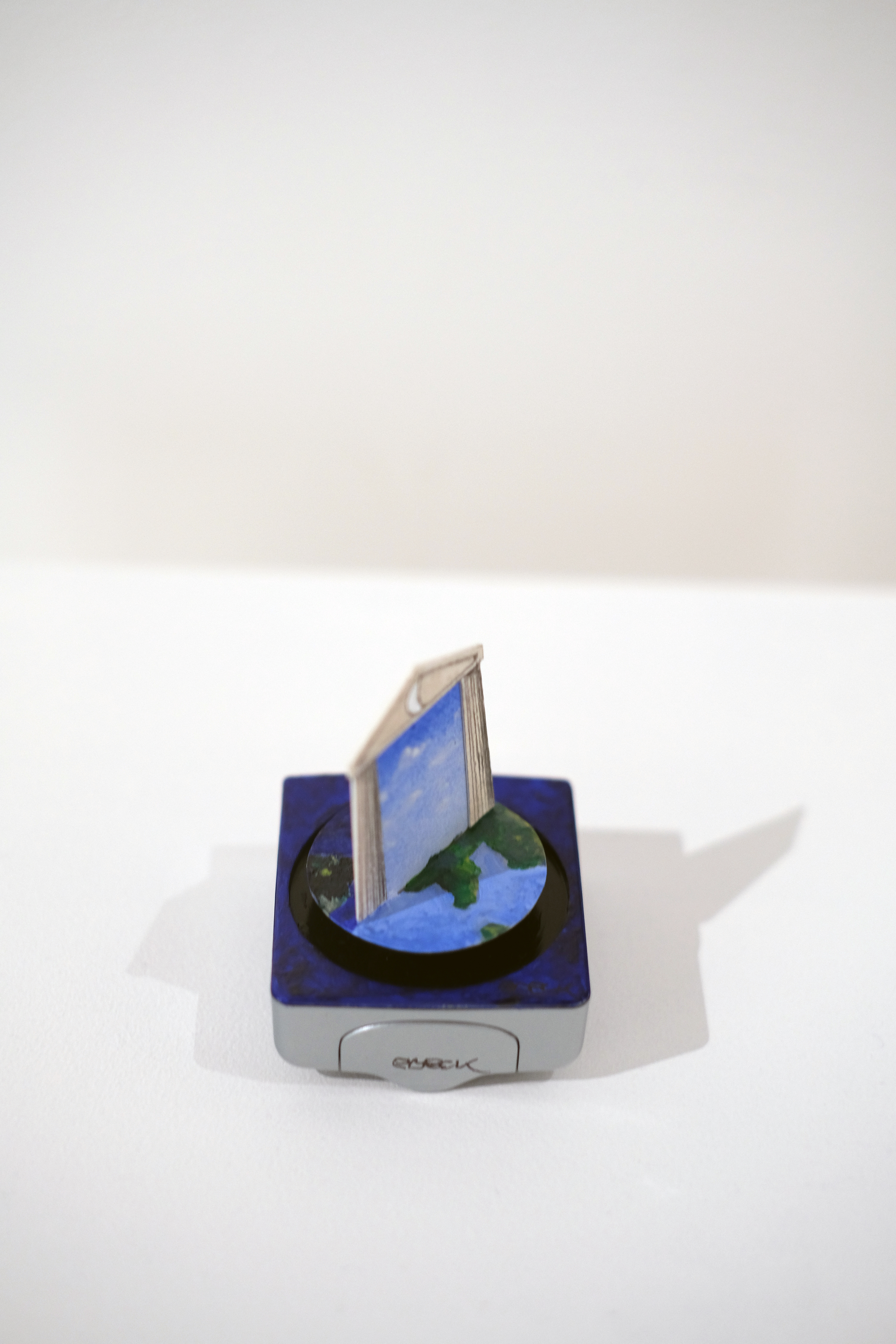 A miniature art installation which rotates on the face of a small battery-powered clock. A paper crafted classical facade depicts blue sky and clouds, set against a pink background, turns to reveal the night time view on the reverse side.