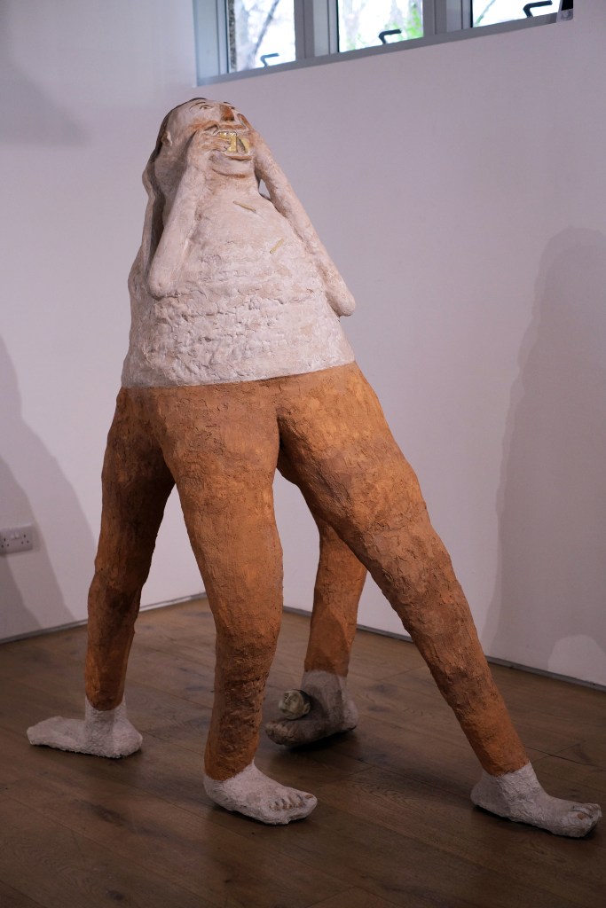 Abstract sculpture of humanoid figure with four elongated orange legs and white, egg-like head, stuffing a yellow object resembling a smartphone into its mouth.