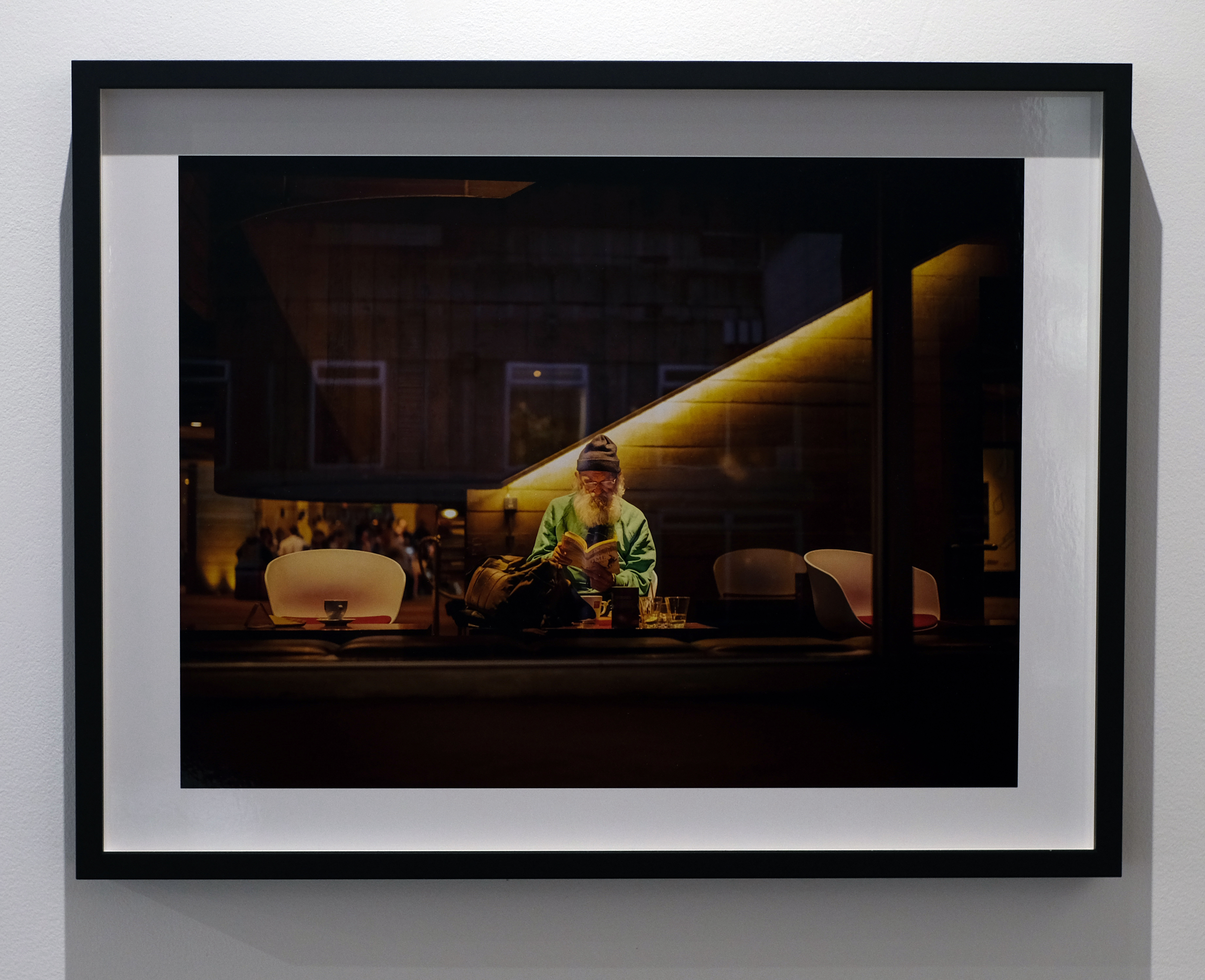 A photograph of an elderly man with a white beard reading a book at a theatre cafe table, with glasses and a backpack beside him, and blurred figures visible in the background.