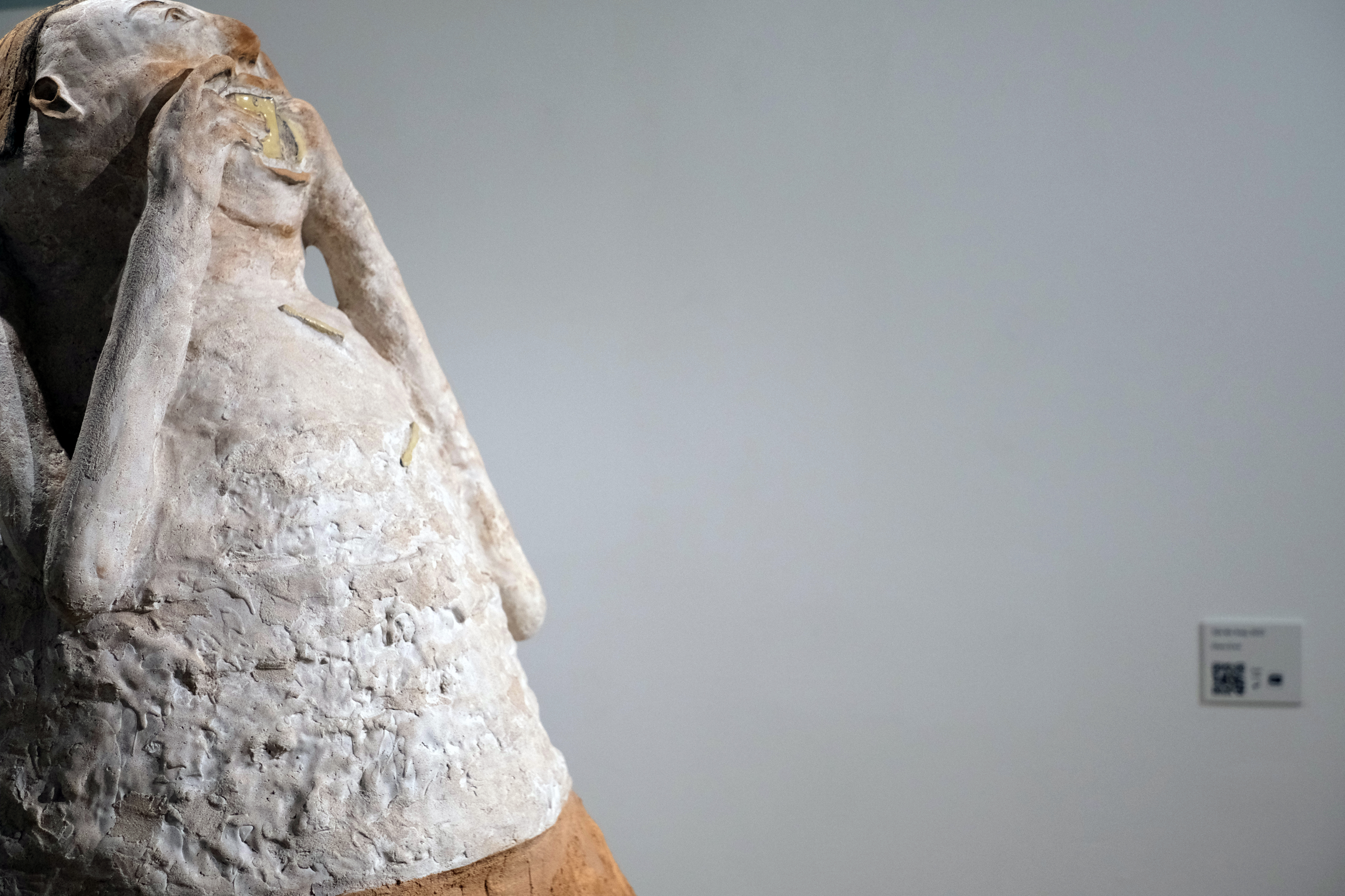 Abstract sculpture of humanoid figure with four elongated orange legs and white, egg-like head, stuffing a yellow object resembling a smartphone into its mouth.