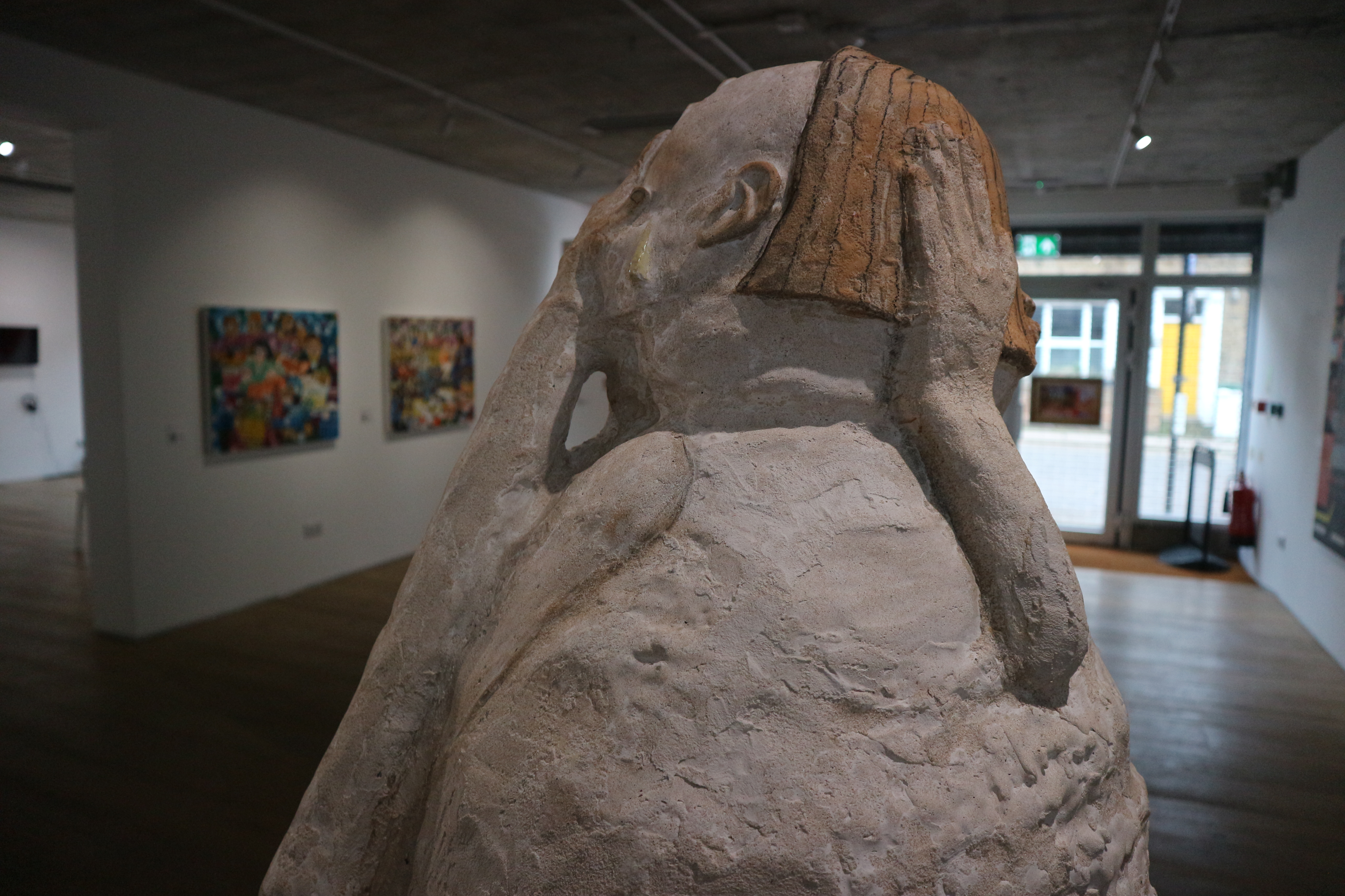 Close up of abstract sculpture of humanoid figure with a white, egg-like head and arms raised photographed from behind.