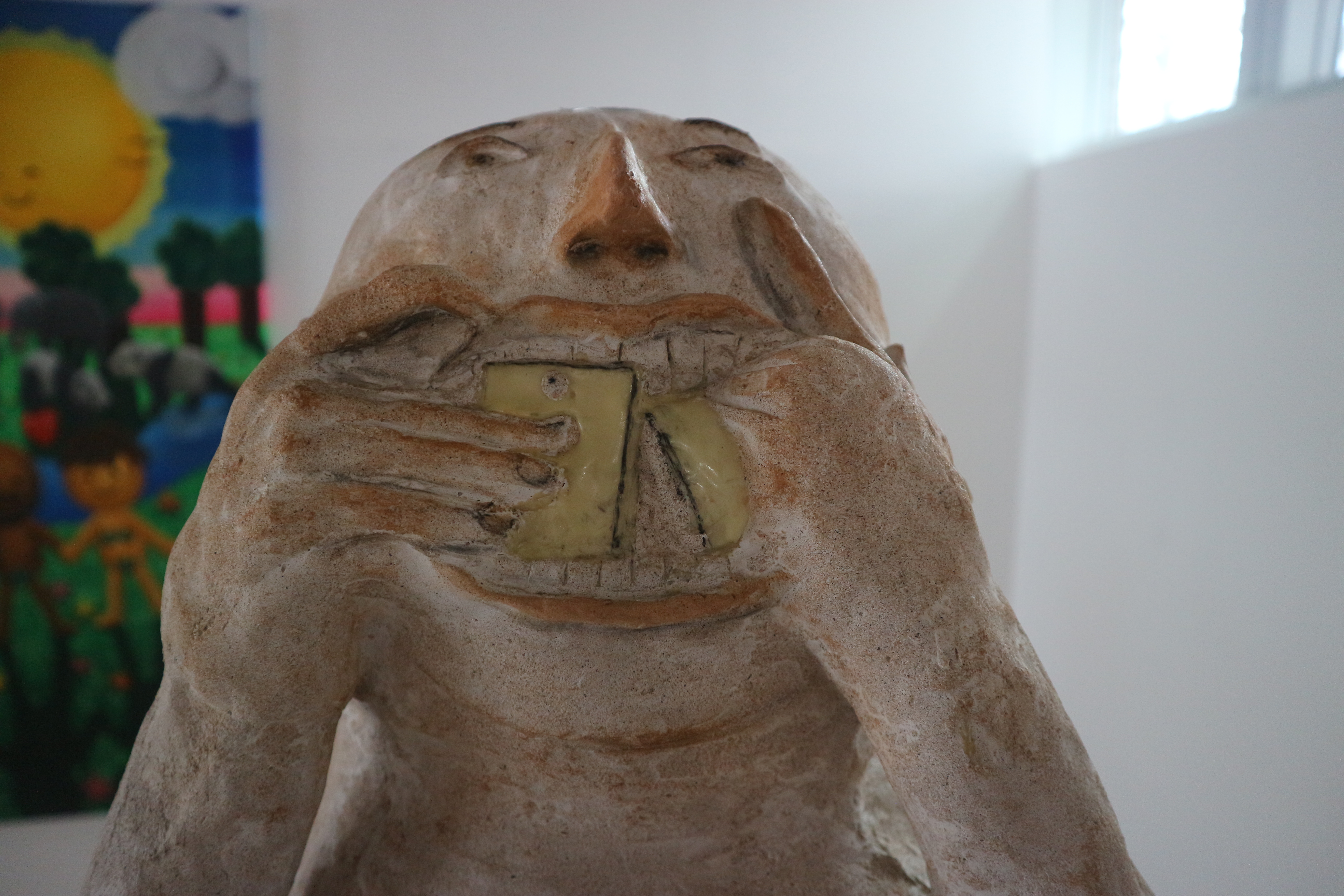 Close up of abstract sculpture of humanoid figure with a white, egg-like head, stuffing a yellow object resembling a smartphone into its mouth.