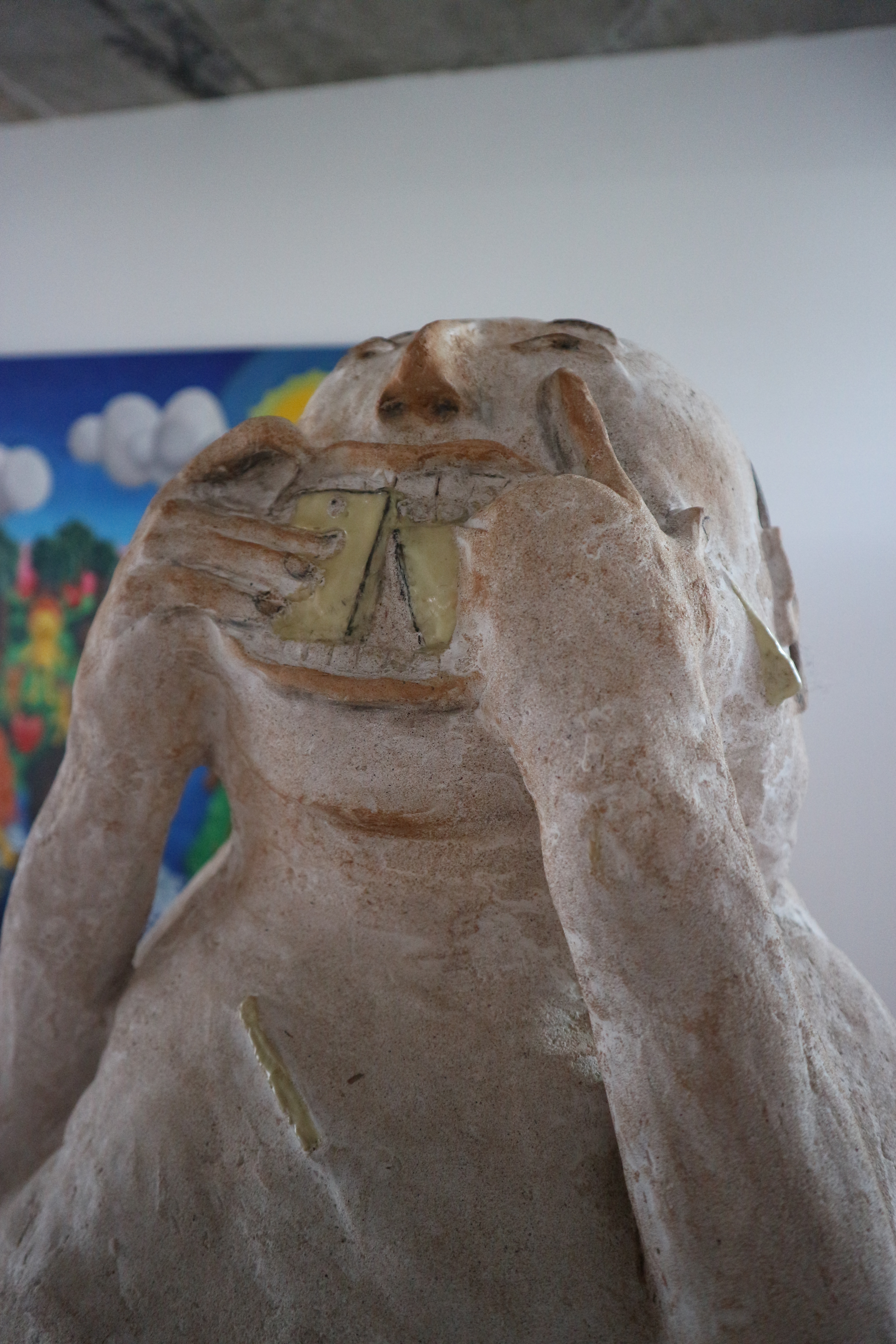 Close up of abstract sculpture of humanoid figure with a white, egg-like head, stuffing a yellow object resembling a smartphone into its mouth.