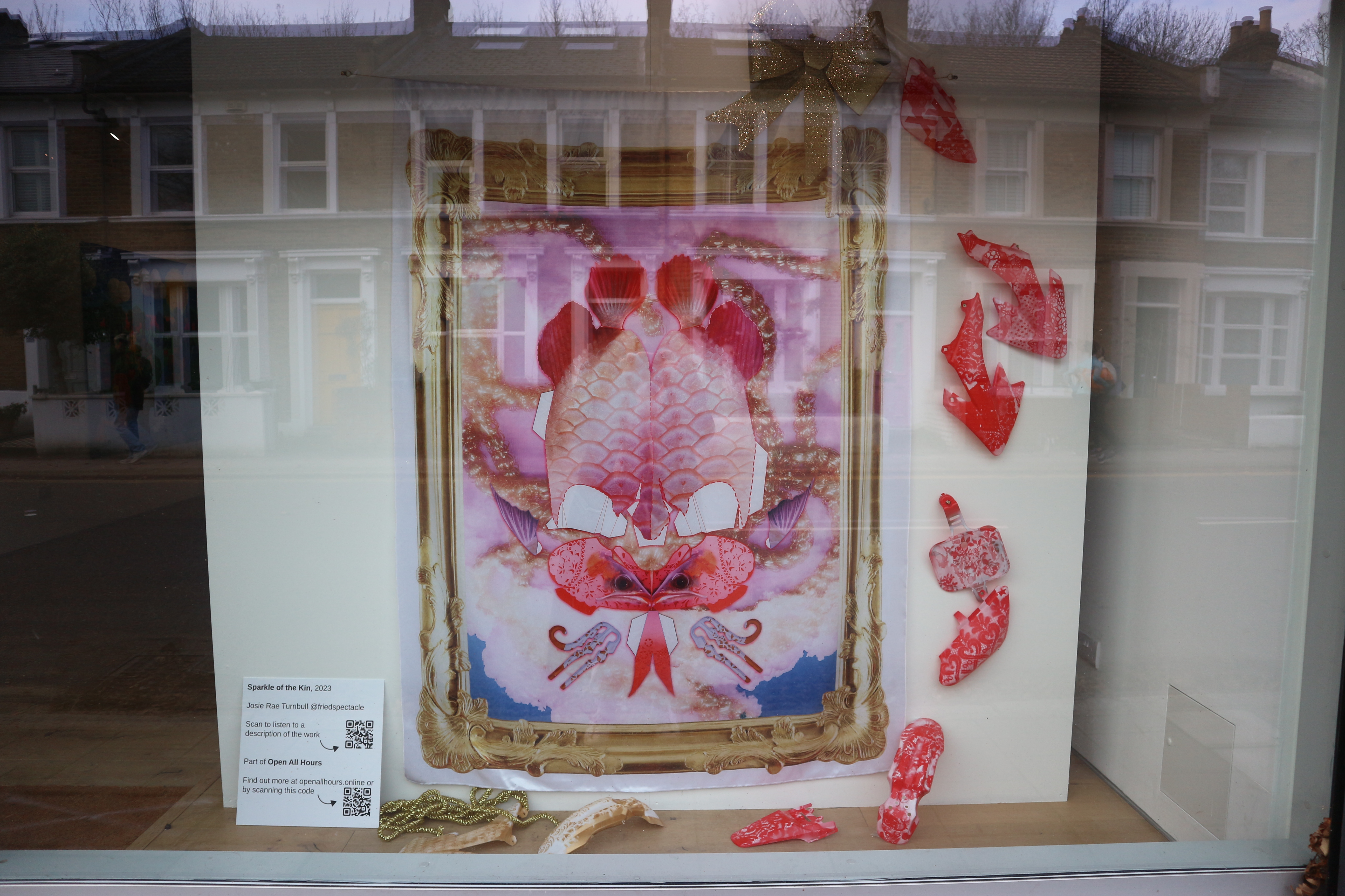 A large digitally printed fabric is hung on a white wall in a large gallery window. It has a printed gold frame and fish scales. Around the fabric are red toy car parts with white spray painted patterns.