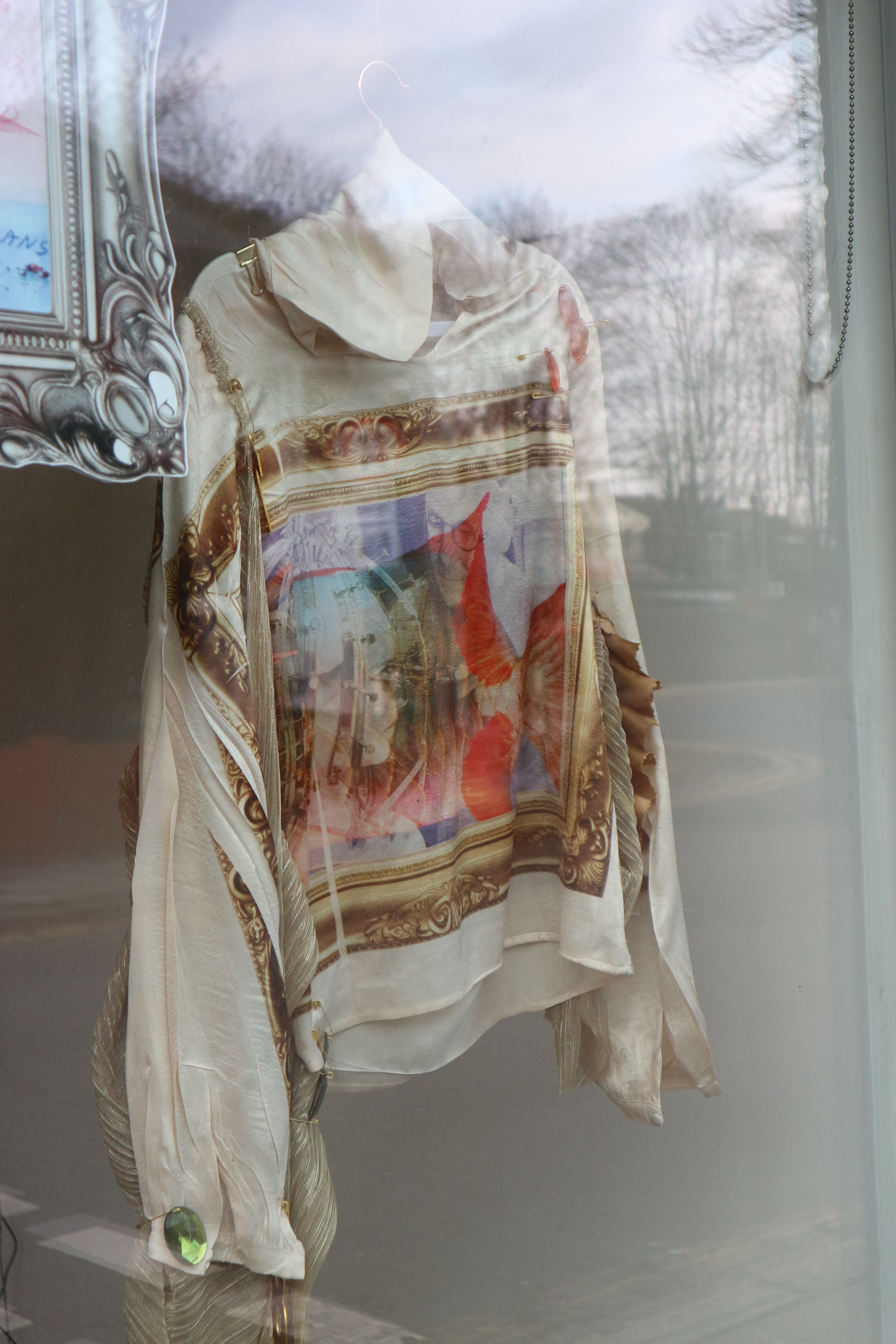 Close up photo of a white costume with a digitally printed design on with resembling a gold frame. Its hung in a gallery window..