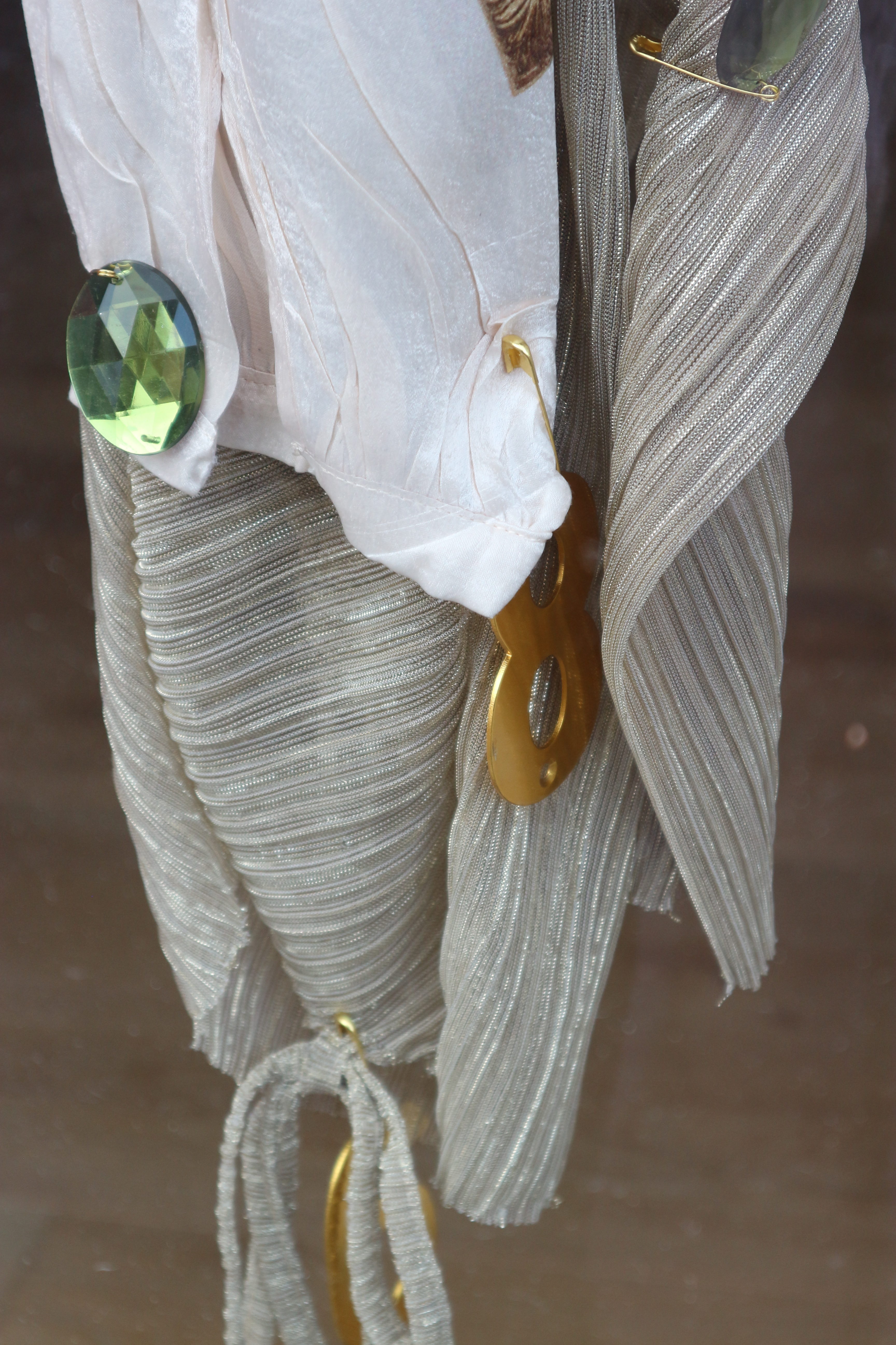 Close up photo of shiny fabric of different textures, with a large green gem attached as well as golden safety pins.