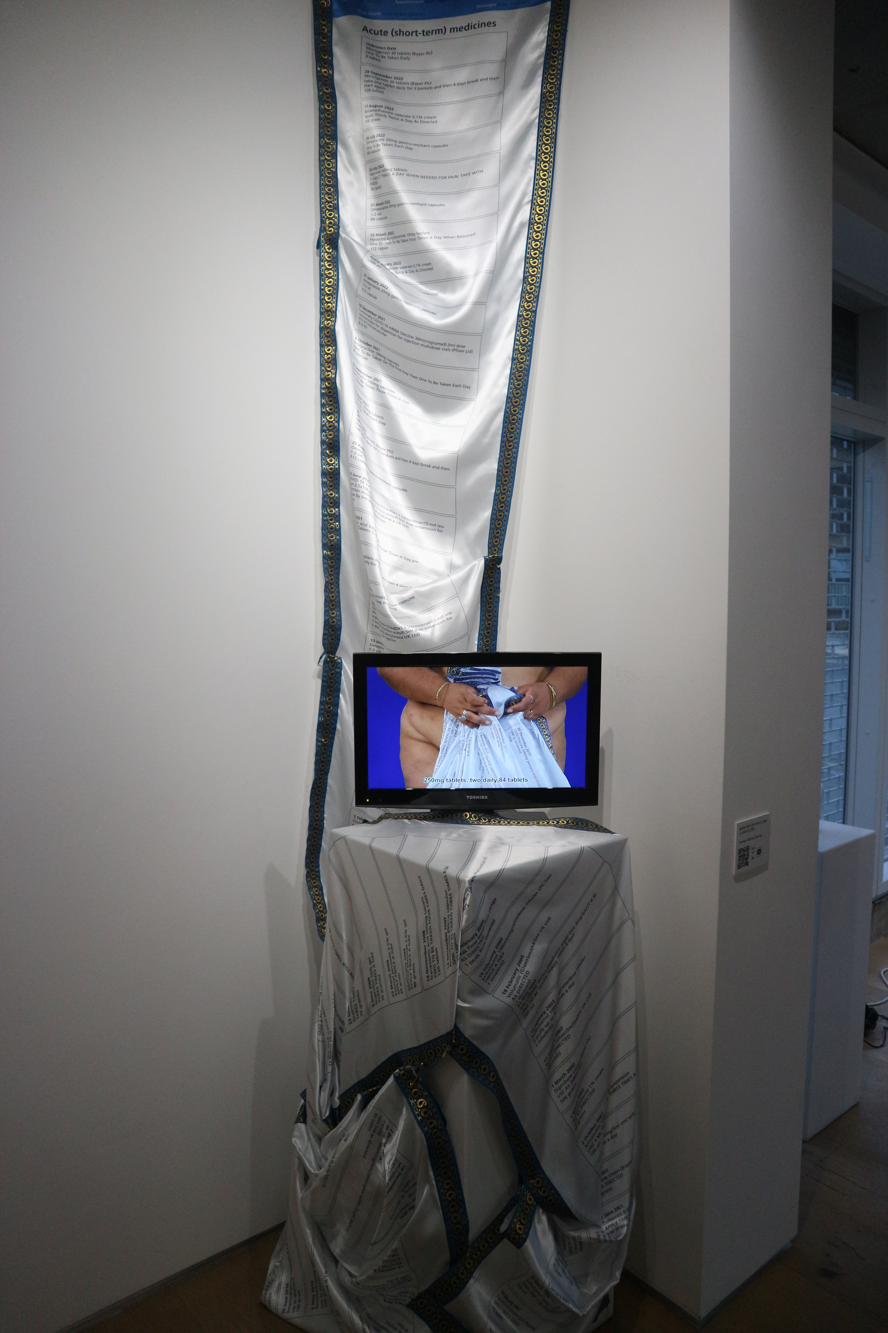 A long roll of silk fabric with prescriptions printed on it hangs down the length of a white wall. In front on a plinth is a small TV screen showing a video of an unclothed woman holding the same fabric.