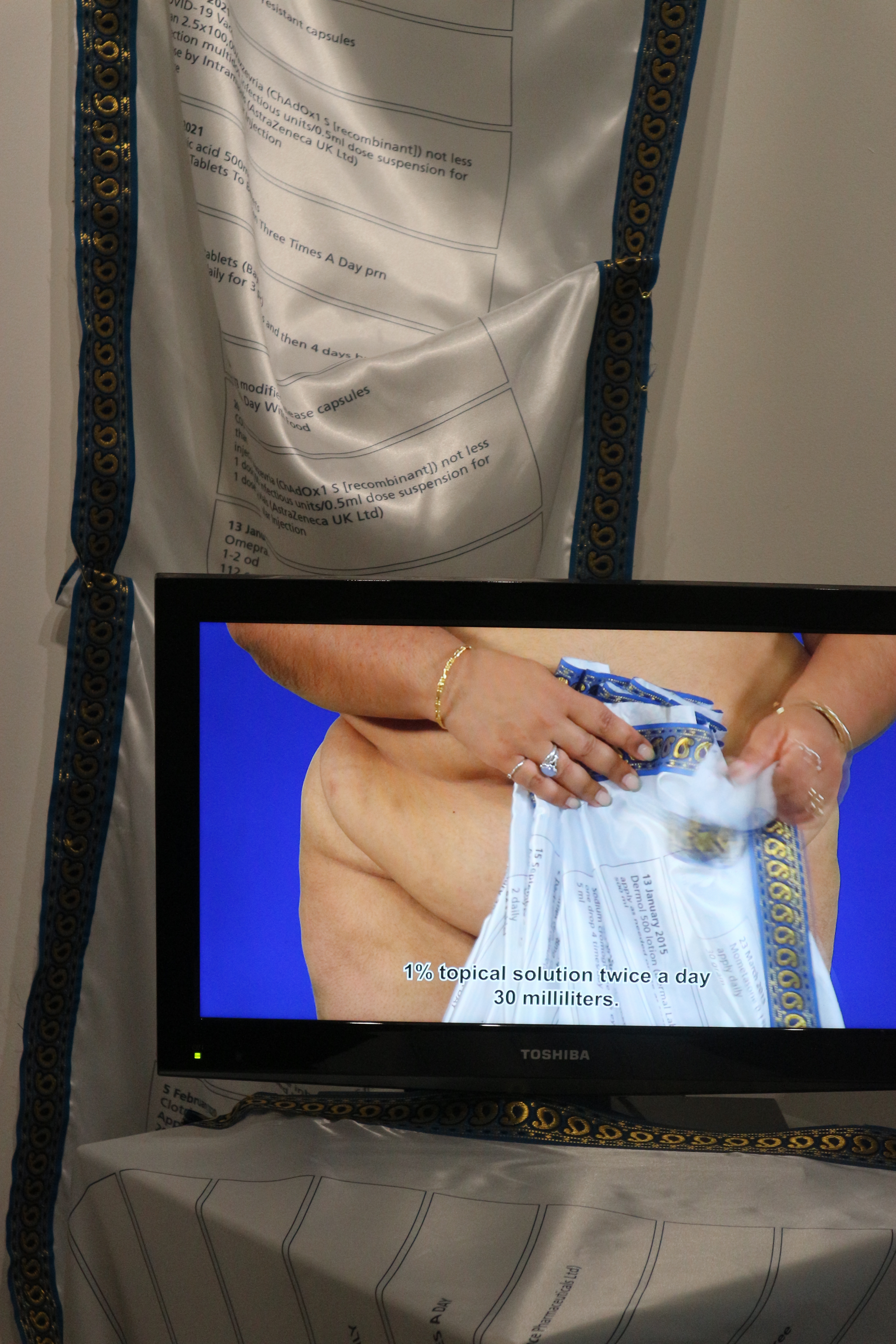 A long roll of silk fabric with prescriptions printed on it hangs down the length of a white wall. In front on a plinth is a small TV screen showing a video of an unclothed woman holding the same fabric.