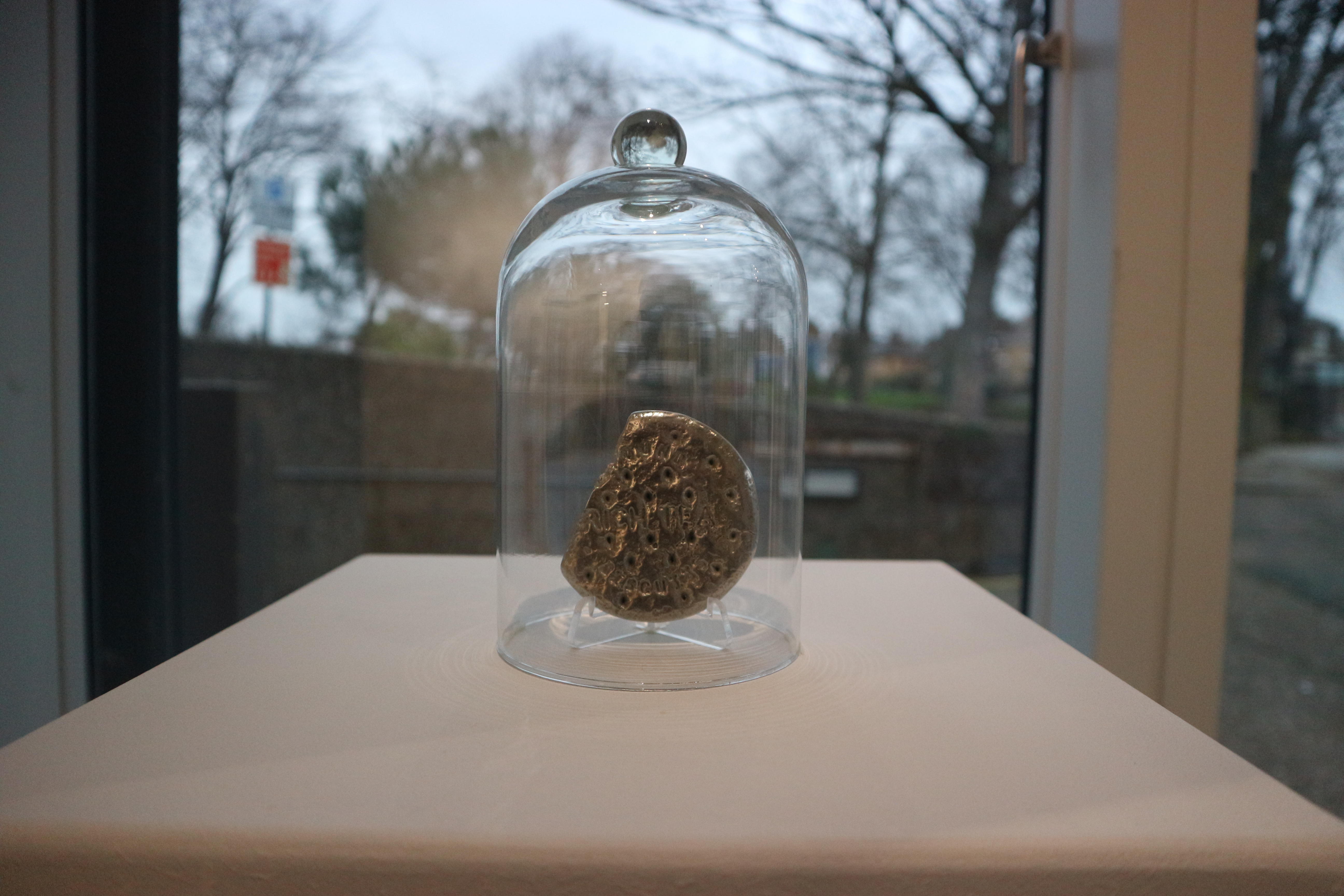 A silver-coloured metal sculpture of a broken round biscuit with the embossed text "OUND RICH TEA," displayed under a clear glass dome on a white base.