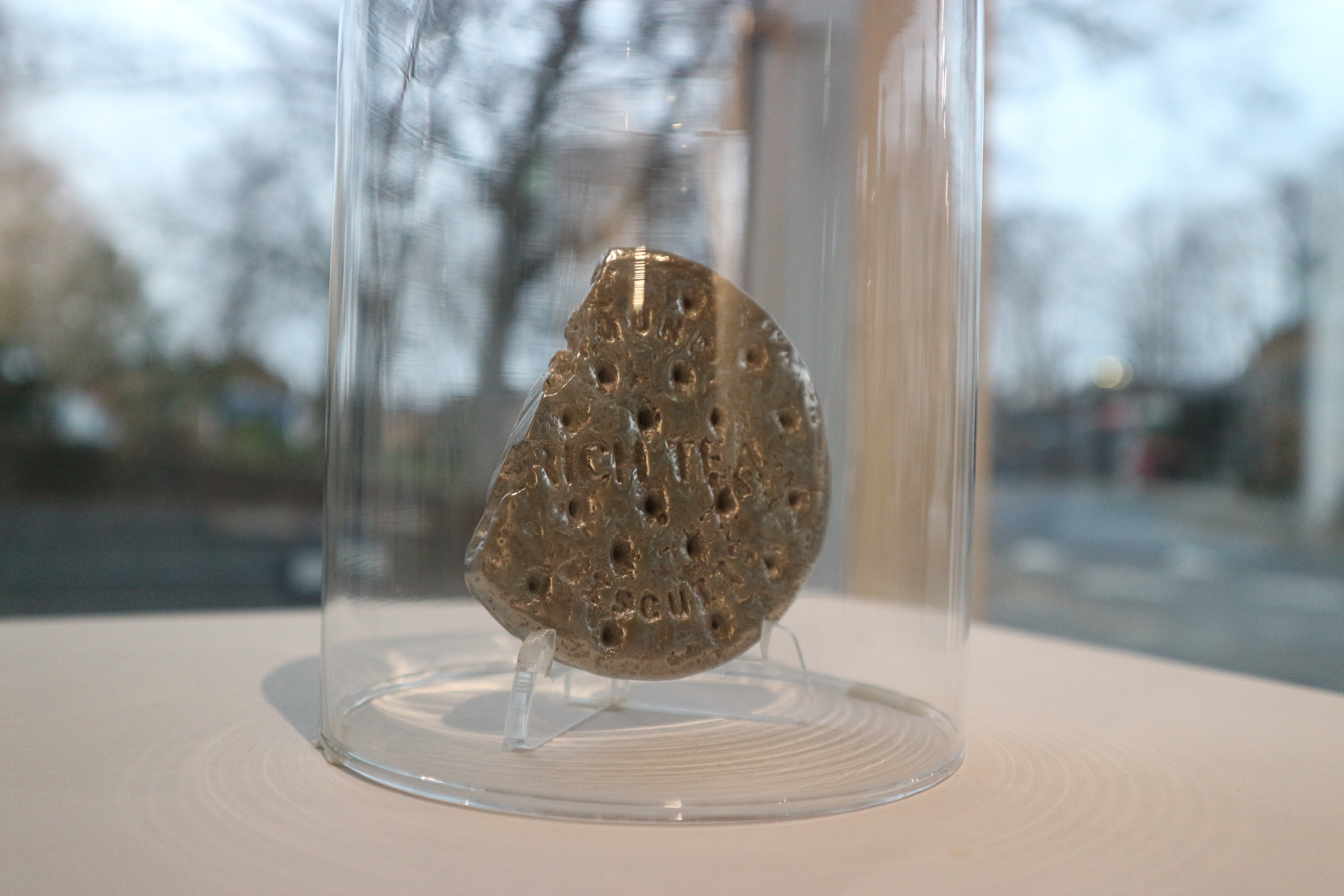 A silver-coloured metal sculpture of a broken round biscuit with the embossed text "OUND RICH TEA," displayed under a clear glass dome on a white base.