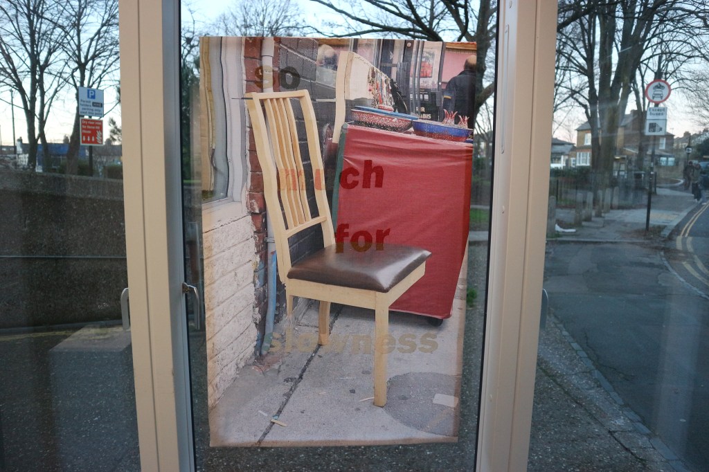 Transparent fabric panel with printed image of side-view two-legged wooden chair and urban street background, with brownish red text reading 'so much for slowness' embroidered into the fabric.