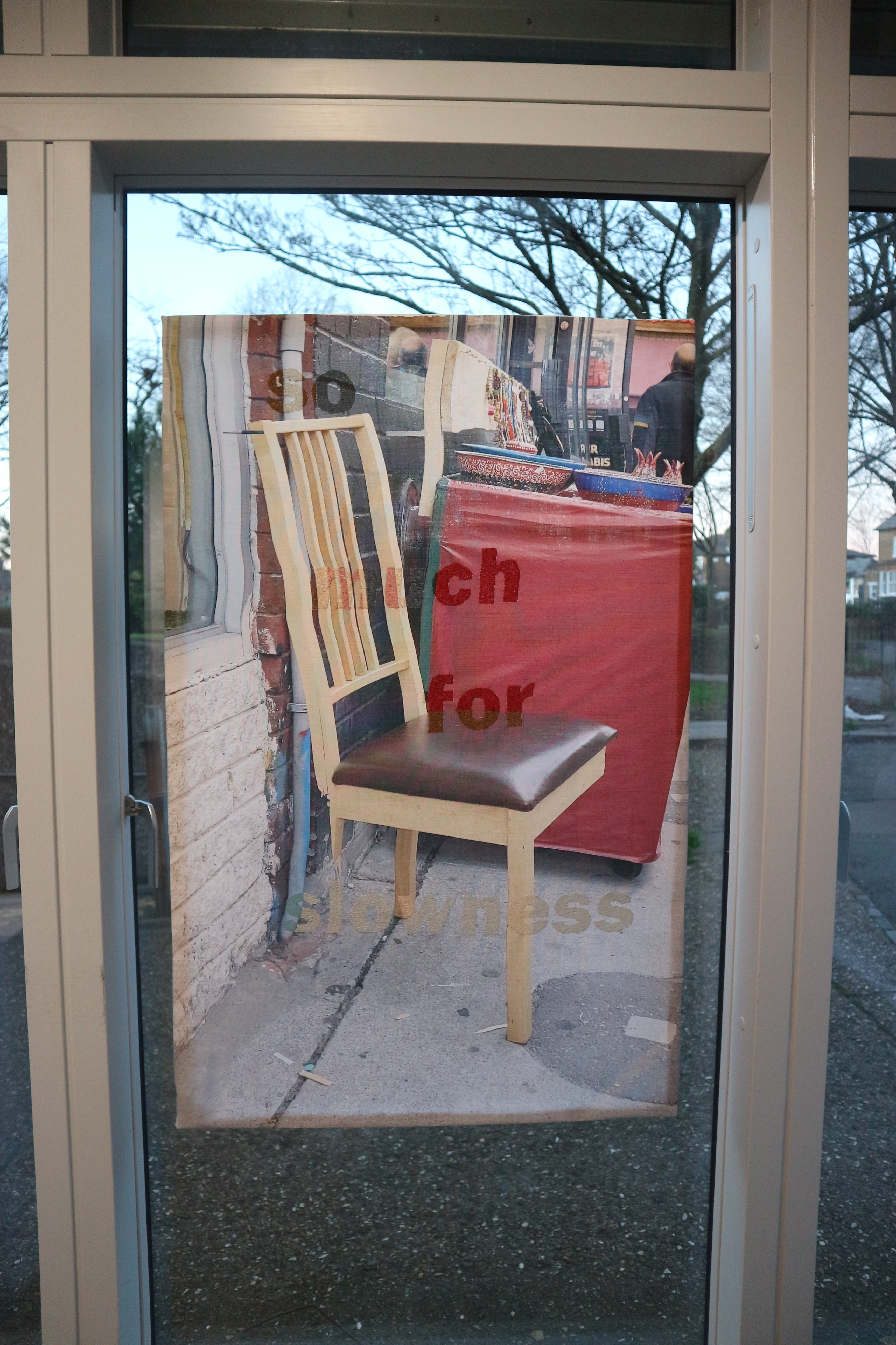 Transparent fabric panel with printed image of side-view two-legged wooden chair and urban street background, with brownish red text reading 'so much for slowness' embroidered into the fabric.