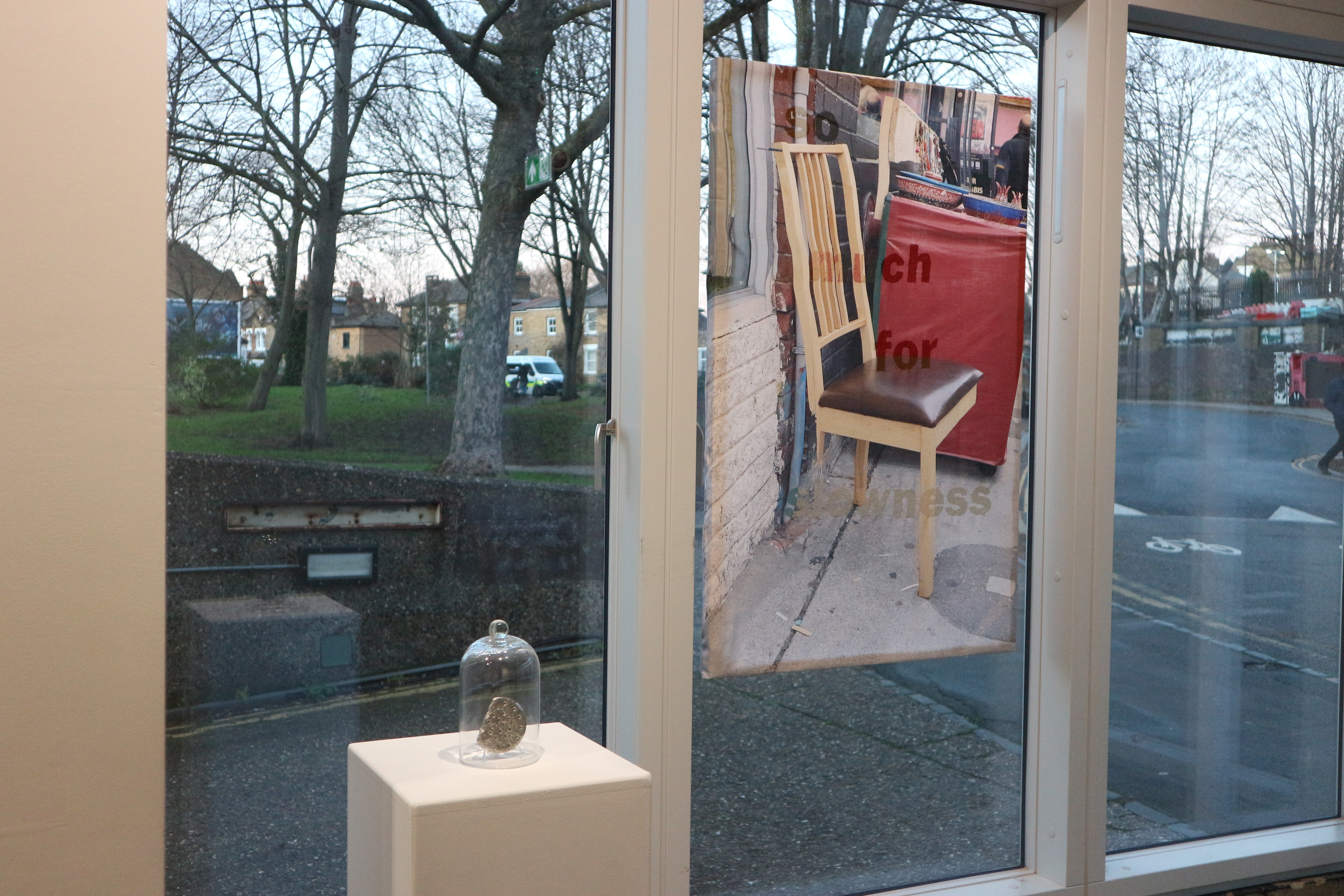 Transparent fabric panel hangs in the gallery window with printed image of side-view two-legged wooden chair and urban street background, with brownish red text reading 'so much for slowness' embroidered into the fabric. To the left is a small sculpture under a glass bell jar on a plinth.